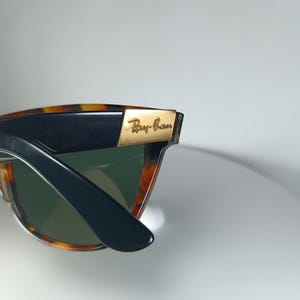 May include: Close-up of Ray-Ban sunglasses. The frames are a tortoiseshell pattern with black arms. The lenses are green. The Ray-Ban logo is visible in gold lettering on the frame.