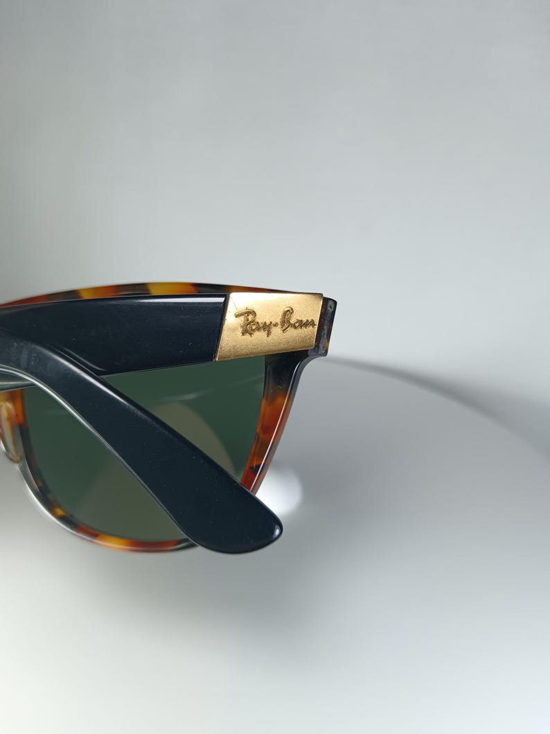 May include: Close-up of Ray-Ban sunglasses. The frames are a tortoiseshell pattern with black arms. The lenses are green. The Ray-Ban logo is visible in gold lettering on the frame.