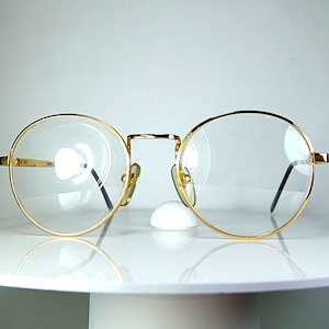 Könnte beinhalten: Goldrandbrille mit runden Gläsern. Die Brille hat ein klassisches Design mit Steg und Bügeln. Die Gläser sind klar und der Rahmen ist goldfarben glänzend. Die Brille wird auf einer weißen Oberfläche präsentiert.