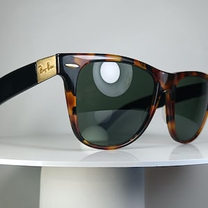 May include: Sunglasses with a classic design. The frames are a tortoiseshell pattern with black arms and dark green lenses. The Ray-Ban logo is visible on the gold-colored metal hinge.