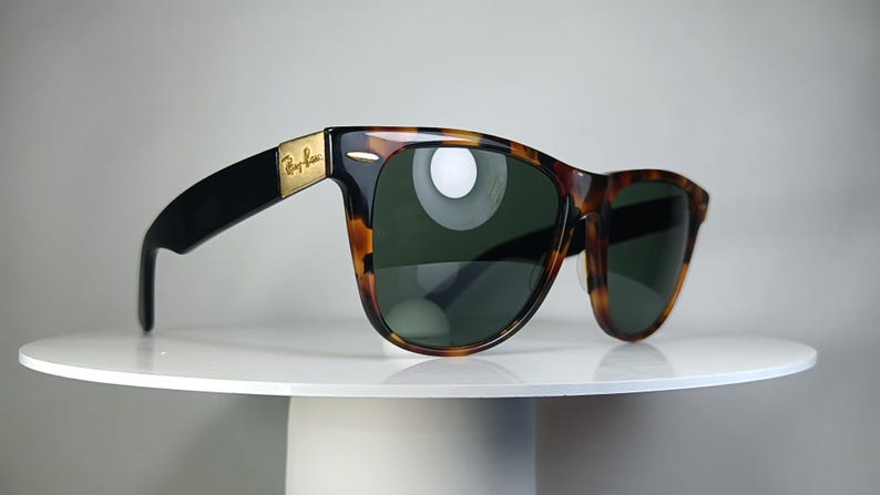 May include: Sunglasses with a classic design. The frames are a tortoiseshell pattern with black arms and dark green lenses. The Ray-Ban logo is visible on the gold-colored metal hinge.