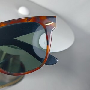 May include: Close-up of sunglasses with a tortoiseshell frame and dark lenses. The sunglasses have a classic design with a gold accent on the frame. The lenses are a dark green color.