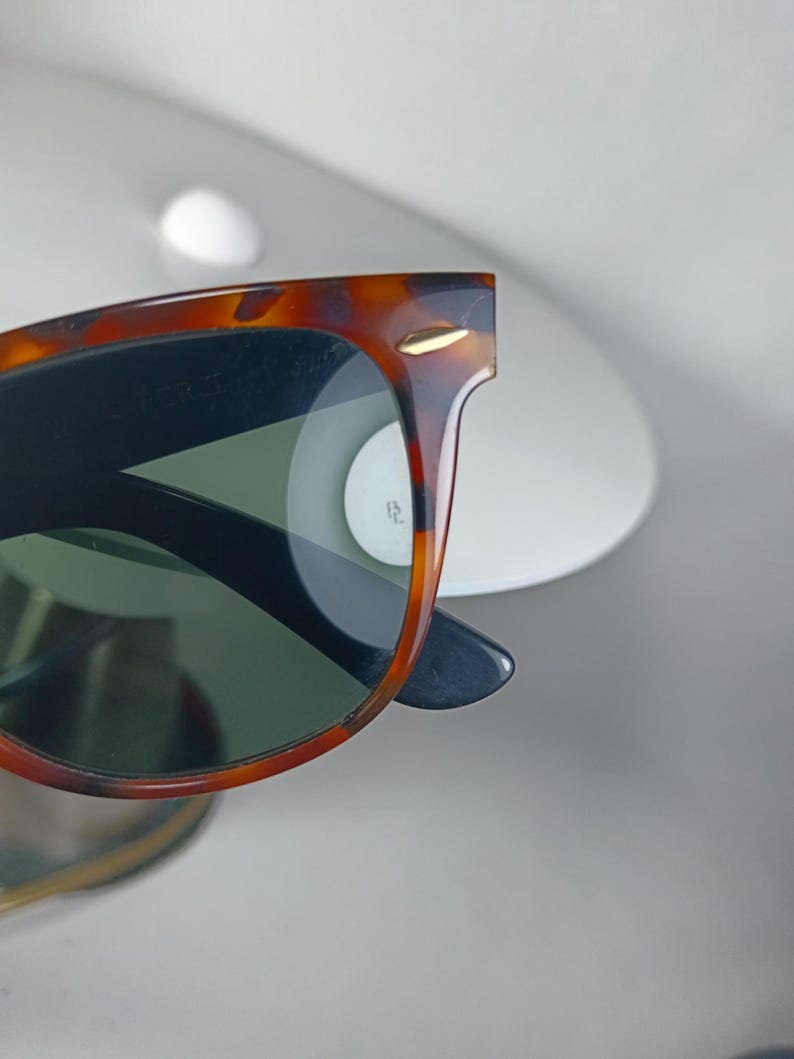 May include: Close-up of sunglasses with a tortoiseshell frame and dark lenses. The sunglasses have a classic design with a gold accent on the frame. The lenses are a dark green color.