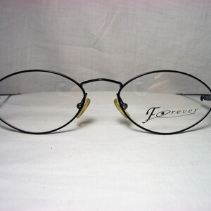 Puede incluir: Gafas ovaladas negras con un fino marco de metal y lentes transparentes. El nombre de la marca "Forever" está impreso en la lente derecha.