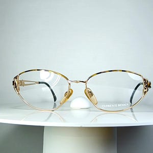 Könnte beinhalten: Goldfarbene Brille mit ovalen Gläsern und schwarzen Bügeln. Die Fassungen haben ein Schildpattmuster an den oberen Rändern und dekorative Akzente. Der Markenname "FLORENCE DESIGN" ist auf dem unteren Rand aufgedruckt.