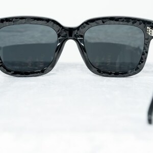 Puede incluir: Gafas de sol negras con montura texturizada y lentes oscuros. Las gafas se muestran desde atr&aacute;s, con las patillas extendidas hacia los lados.