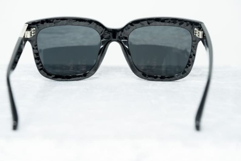 Puede incluir: Gafas de sol negras con montura texturizada y lentes oscuros. Las gafas se muestran desde atr&aacute;s, con las patillas extendidas hacia los lados.