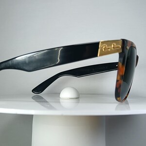May include: Black sunglasses with a tortoiseshell pattern on the frames and lenses. The sunglasses have a gold accent on the temples and are resting on a white surface. The sunglasses are a classic style.