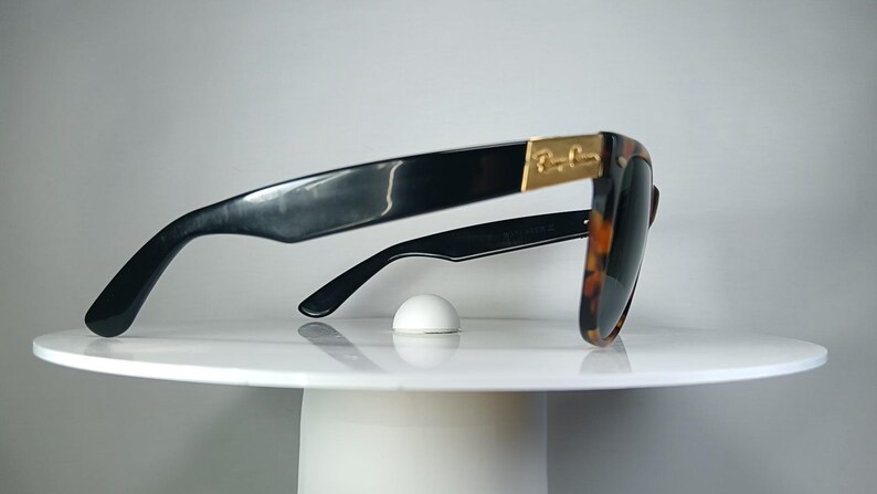 May include: Black sunglasses with a tortoiseshell pattern on the frames and lenses. The sunglasses have a gold accent on the temples and are resting on a white surface. The sunglasses are a classic style.