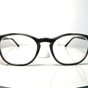 May include: Black-framed eyeglasses with a round shape, resting on a white surface. The glasses have clear lenses and a subtle, dark gray hue. The design is simple and classic, suitable for everyday wear.
