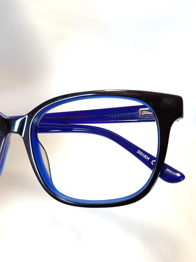 May include: Close-up of eyeglasses with a black frame and blue interior. The arms and lenses are blue. The text "5001804 C" is printed on the arm of the glasses.