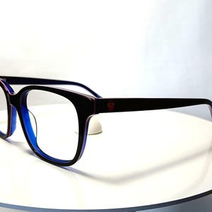 May include: Black-framed eyeglasses with a square shape and blue interior. The temples are dark with a red accent near the hinge. The glasses are set on a white surface.