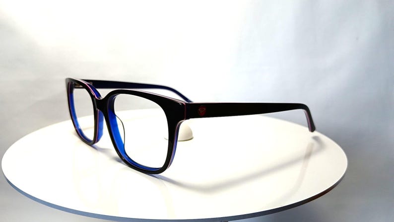 May include: Black-framed eyeglasses with a square shape and blue interior. The temples are dark with a red accent near the hinge. The glasses are set on a white surface.