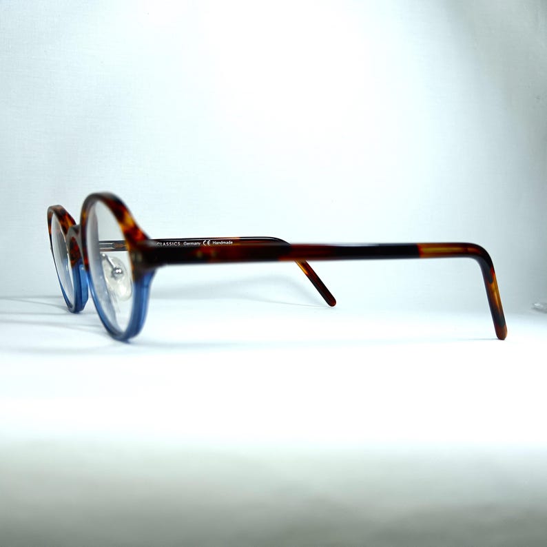 May include: Eyeglasses with a tortoiseshell pattern on the upper frames and temples, transitioning to a blue hue on the lower frames. The glasses have a classic, round shape and are set against a white background.