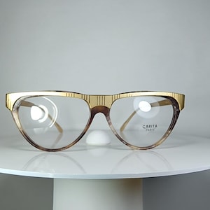 Könnte beinhalten: Eine Vintage-Brille mit goldfarbenem Rahmen und klaren Gläsern. Die Oberseite des Rahmens hat ein strukturiertes Design. Die Gläser sind tropfenförmig. Der Markenname "CARITA PARIS" ist auf dem rechten Glas aufgedruckt.