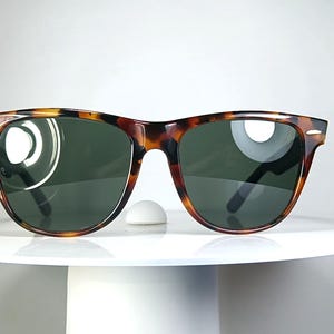 May include: Tortoiseshell sunglasses with dark green lenses. The sunglasses have a classic, rectangular frame design. The glasses are displayed on a white, round pedestal.
