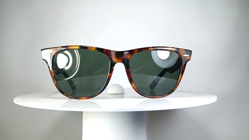May include: Tortoiseshell sunglasses with dark green lenses. The sunglasses have a classic, rectangular frame design. The glasses are displayed on a white, round pedestal.