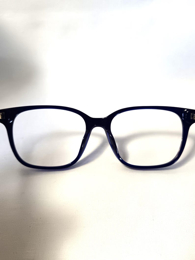 May include: A pair of dark blue rectangular eyeglasses with clear lenses. The frames are thick and have a rounded shape at the top corners. The glasses are set against a white background.
