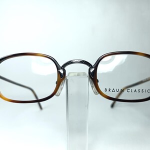 May include: Brown and black Braun Classics eyeglasses with a tortoiseshell pattern on the frames. The glasses have a metal bridge and thin temples. The brand name is printed on the right lens.