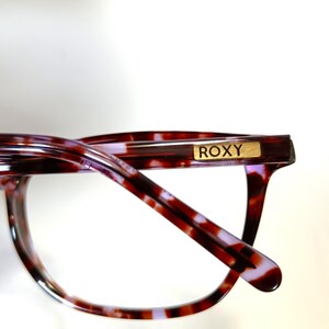 Peut inclure: Gros plan de lunettes avec un motif &eacute;caille de tortue dans les tons marron, rouge et violet. La monture est de forme rectangulaire et porte le nom de la marque "ROXY" en lettres dor&eacute;es sur la branche.