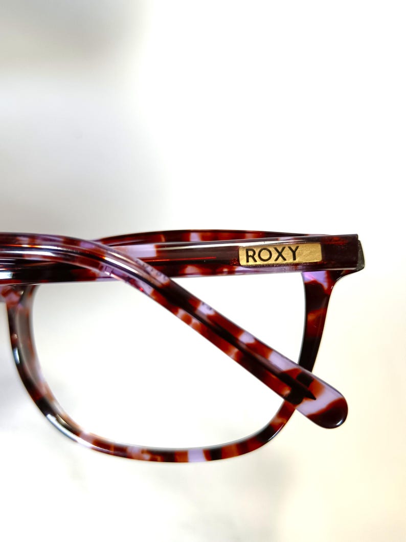 Peut inclure: Gros plan de lunettes avec un motif &eacute;caille de tortue dans les tons marron, rouge et violet. La monture est de forme rectangulaire et porte le nom de la marque "ROXY" en lettres dor&eacute;es sur la branche.