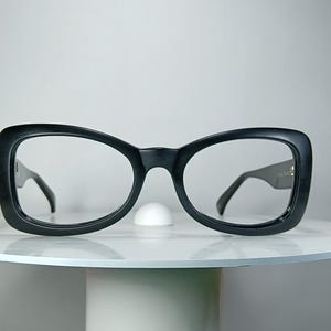 Könnte beinhalten: Eine schwarze Brille mit dicken, abgerundet rechteckigen Rahmen und klaren Gläsern. Die Brille verfügt über einen markanten Steg und breite Bügel, die nach außen verlaufen, und besticht durch eine durchgehend kräftige, schwarze Farbgebung.
