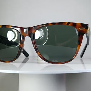 May include: Tortoiseshell sunglasses with dark green lenses. The frames have a classic design with a brown and black pattern. The sunglasses are displayed on a white, round pedestal.
