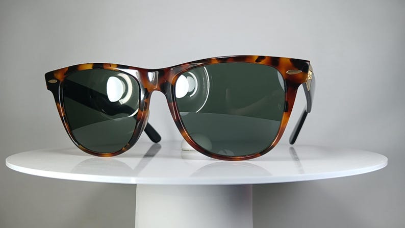 May include: Tortoiseshell sunglasses with dark green lenses. The frames have a classic design with a brown and black pattern. The sunglasses are displayed on a white, round pedestal.