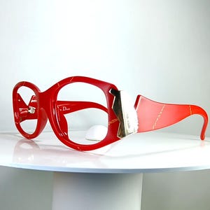 May include: Bright red oversized eyeglasses featuring a thick glossy frame with a rounded shape. The temples are accented with a prominent gold-tone metal hinge detail on each side, and the inner temple arm displays the Christian Dior brand name.
