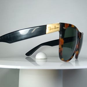 May include: Classic Ray-Ban sunglasses with a tortoiseshell pattern frame and dark lenses. The temples are black, and the Ray-Ban logo is visible on the gold-colored metal accents. The sunglasses are resting on a white surface.