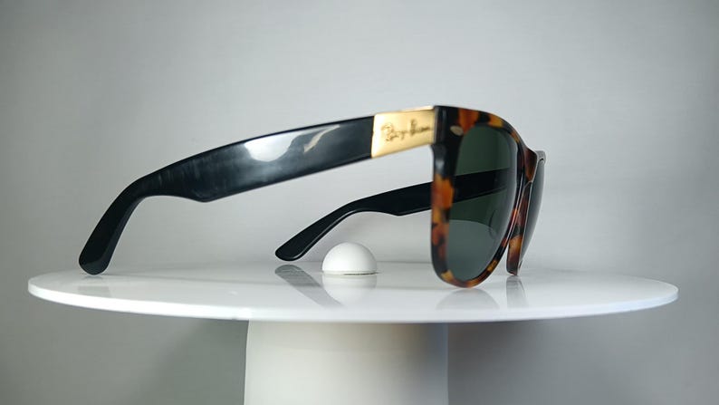 May include: Classic Ray-Ban sunglasses with a tortoiseshell pattern frame and dark lenses. The temples are black, and the Ray-Ban logo is visible on the gold-colored metal accents. The sunglasses are resting on a white surface.