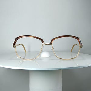 Puede incluir: Gafas con una montura de metal fino en tono dorado y un patrón de carey a lo largo de la línea superior. Las lentes son transparentes y rectangulares con bordes redondeados, sostenidas por un puente fino y varillas delgadas a los lados.