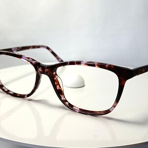 Peut inclure: Lunettes &agrave; motif &eacute;caille de tortue. Les montures sont un m&eacute;lange de marron, de noir et de blanc, de forme rectangulaire et avec des verres transparents. Les lunettes reposent sur une surface blanche.