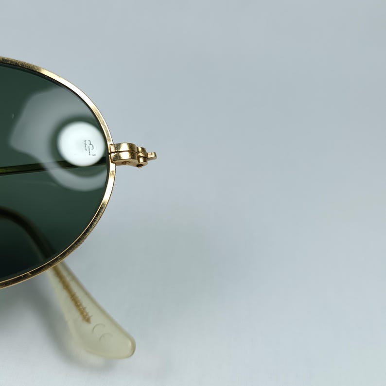 Puede incluir: Gafas de sol aviador doradas con lentes verdes. Las gafas tienen un marco de metal y un puente nasal de pl&aacute;stico transparente. El nombre de la marca "B&L" es visible en el marco.