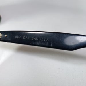 May include: Close-up of a black Ray-Ban eyeglass arm. The text "B&L RAY-BAN U.S.A." is printed in white on the arm. The arm is angled, and the background is a plain white surface.