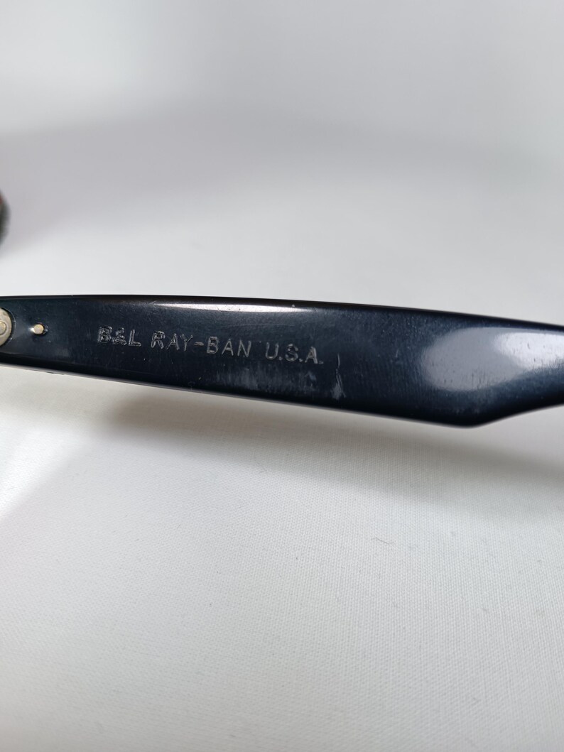May include: Close-up of a black Ray-Ban eyeglass arm. The text "B&L RAY-BAN U.S.A." is printed in white on the arm. The arm is angled, and the background is a plain white surface.
