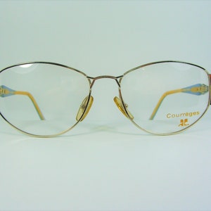 May include: Gold-tone metal eyeglasses with a cat-eye shape. The frames have a red, green, and blue design on the temples. The brand name "Courr&egrave;ges" is printed on the right temple.