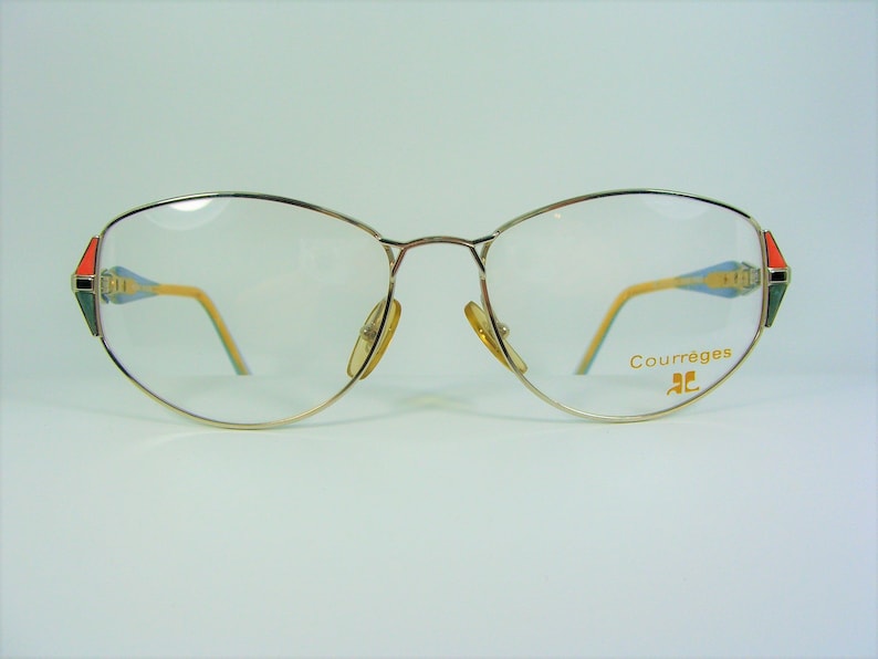 May include: Gold-tone metal eyeglasses with a cat-eye shape. The frames have a red, green, and blue design on the temples. The brand name "Courr&egrave;ges" is printed on the right temple.