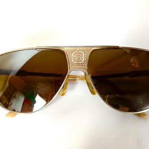 May include: Gold-toned aviator sunglasses with brown lenses. The bridge features a decorative emblem. The frames have a classic design with a double bridge and curved temples. The lenses are reflective.