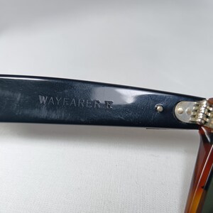 May include: Close-up of a black Wayfarer II sunglasses arm. The text "WAYFARER II" is printed in white on the black arm. The frame is black with a tortoise shell pattern on the arm.
