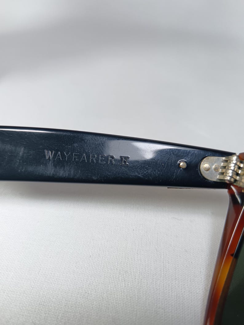 May include: Close-up of a black Wayfarer II sunglasses arm. The text "WAYFARER II" is printed in white on the black arm. The frame is black with a tortoise shell pattern on the arm.