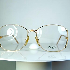 Könnte beinhalten: Goldgerahmte Brille mit klaren Gläsern. Die Brille hat eine rechteckige Form mit abgerundeten Kanten und einem Doppelsteg. Der Markenname "L'AMY PARIS" ist auf dem rechten Glas sichtbar. Die Brille wird auf einer weißen Oberfläche präsentiert.