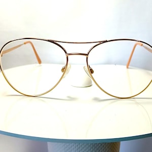 Puede incluir: Gafas de aviador doradas con lentes transparentes. Las gafas tienen un doble puente y una forma clásica de lágrima. Las monturas son de color oro cálido y las patillas son de color melocotón claro. Las gafas se exhiben sobre una superficie blanca.