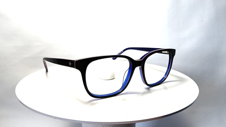 May include: Black eyeglasses with a rectangular frame and blue accents. The glasses have a sleek design with a dark frame and blue interior. The lenses are clear, and the glasses are displayed on a white surface.