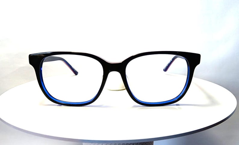 May include: Black-framed eyeglasses with a blue inner rim. The glasses have a square shape and are set against a white background. The temples are thin and have a purple hue.