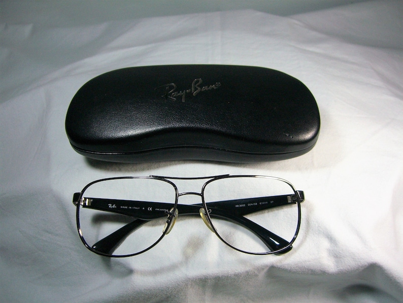 K&ouml;nnte beinhalten: Eine Brille mit silberfarbenem Rahmen, schwarzen B&uuml;geln und klaren Gl&auml;sern. Die Brille liegt auf einer wei&szlig;en Oberfl&auml;che neben einem schwarzen Ray-Ban Brillenetui.