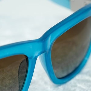 K&ouml;nnte beinhalten: Eine blaue Sonnenbrille mit braunen Gl&auml;sern. Die Sonnenbrille hat einen quadratischen Rahmen und ein elegantes Design.