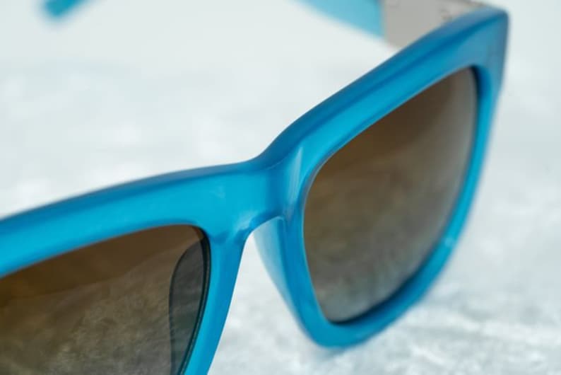 K&ouml;nnte beinhalten: Eine blaue Sonnenbrille mit braunen Gl&auml;sern. Die Sonnenbrille hat einen quadratischen Rahmen und ein elegantes Design.