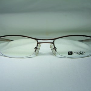 May include: A pair of brown metal eyeglasses with a half-rim design. The lenses are clear and the frames have a subtle curve. The brand name "x-optix the collection" is printed on the right lens.