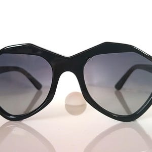 Könnte beinhalten: Schwarze Sonnenbrille mit einem einzigartigen geometrischen Rahmen. Die Gläser haben einen Grauton und die Sonnenbrille ist vor einem weißen Hintergrund platziert. Die Fassungen haben eine markante, eckige Form.